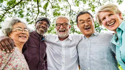 A group of seniors outdoors, standing and smiling in a circle with their arms around each other.