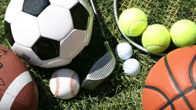 Sports balls placed on the grass. Including a soccer ball, football, baseball, basketball, golf balls and tennis balls, a racquet and a golf club.